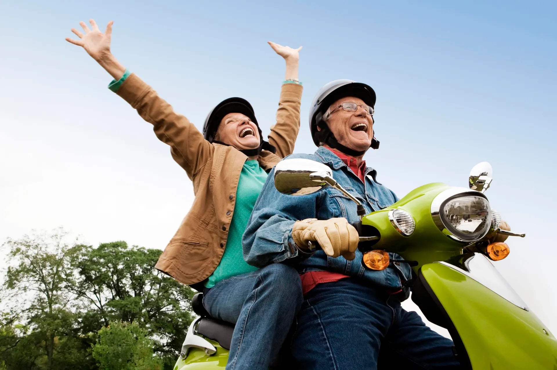 Two people riding a green scooter with arms raised against a clear blue sky.