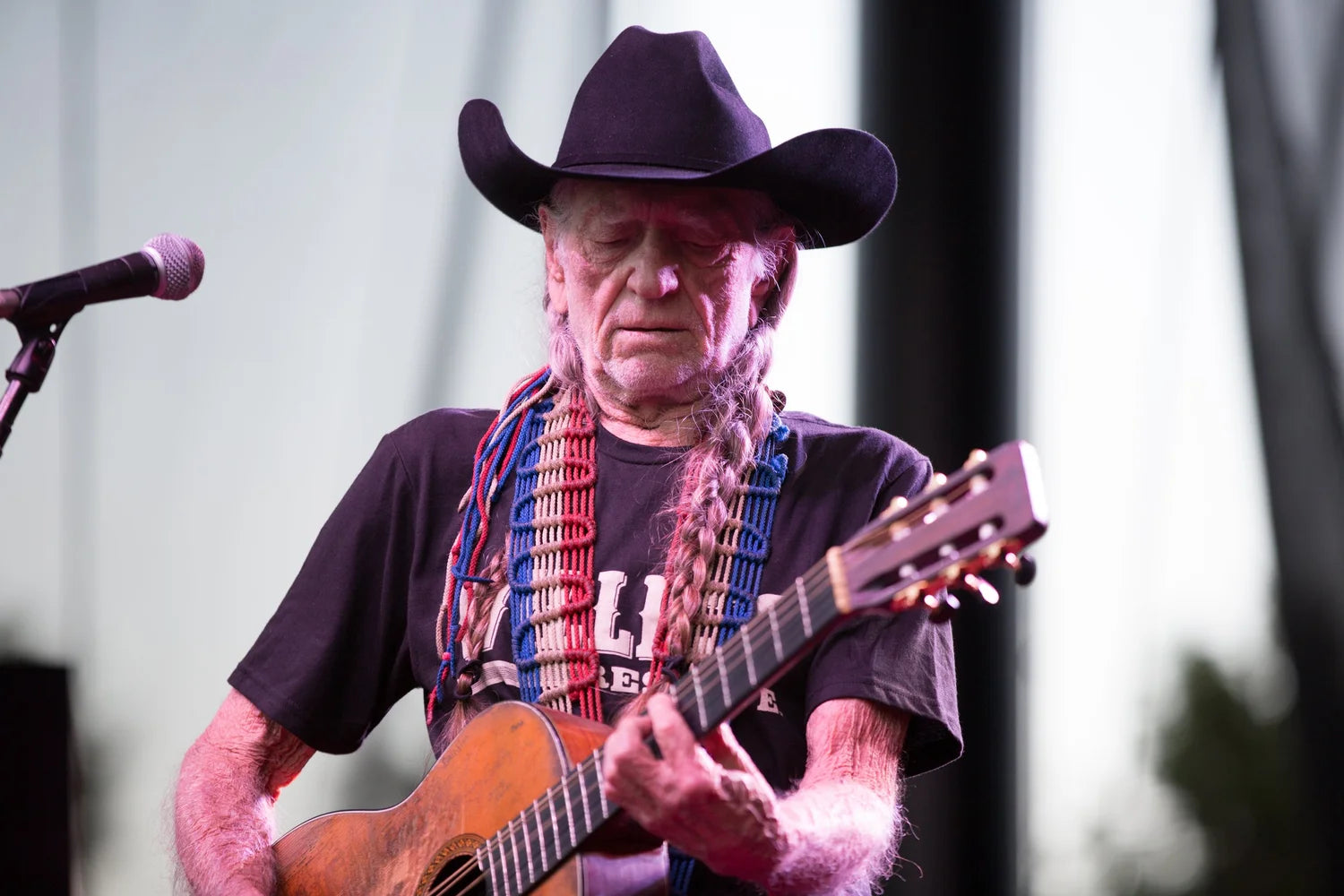 Willie Nelson playing guitar and singing into a microphone with a cowboy hat on.