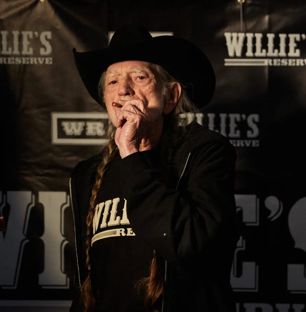 Willie Nelson wearing a black cowboy hat and jacket with 'Willie's Reserve' branding in a promotional setting.