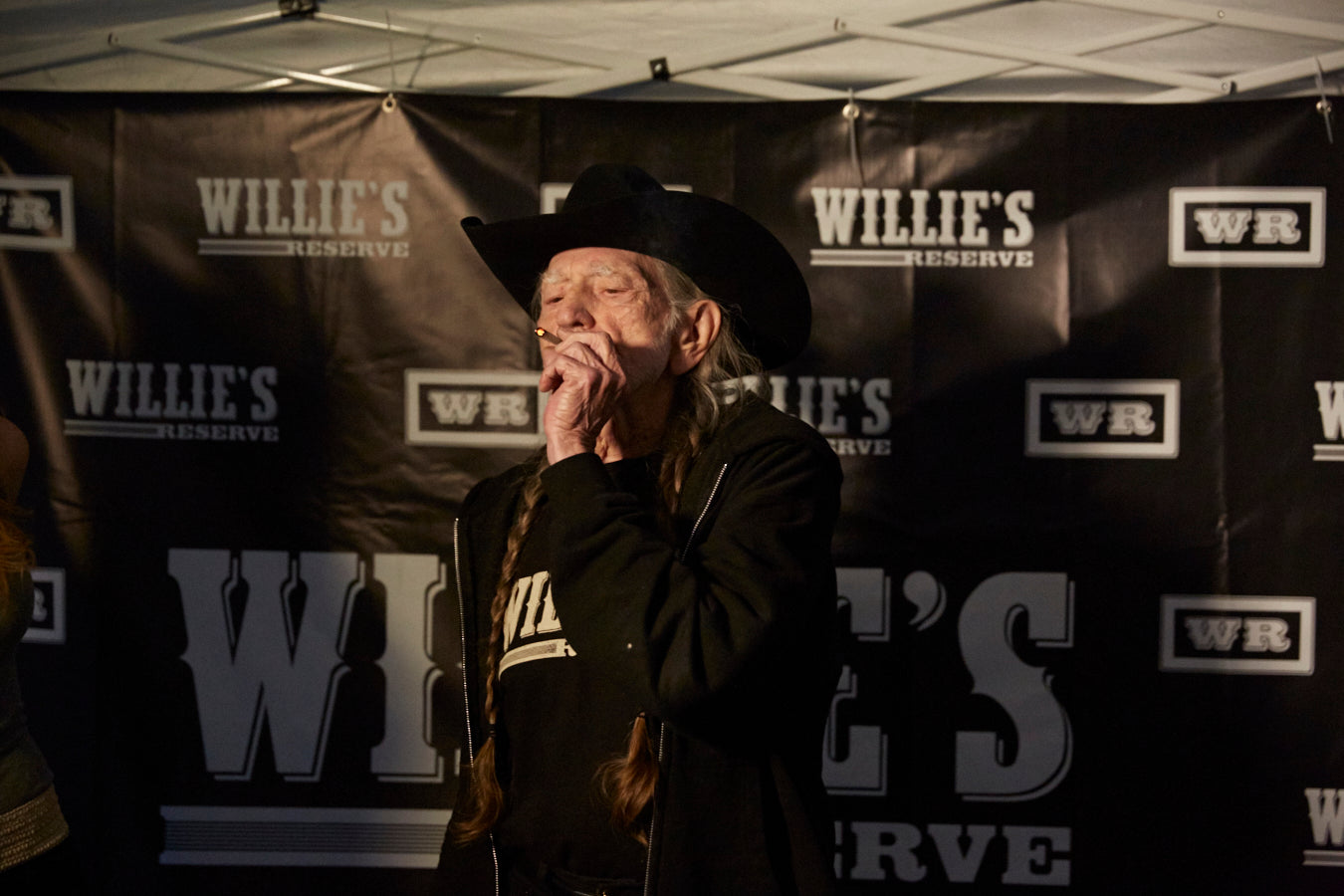 Willie Nelson in a cowboy hat and black coat standing in front of Willies Reserve banners.