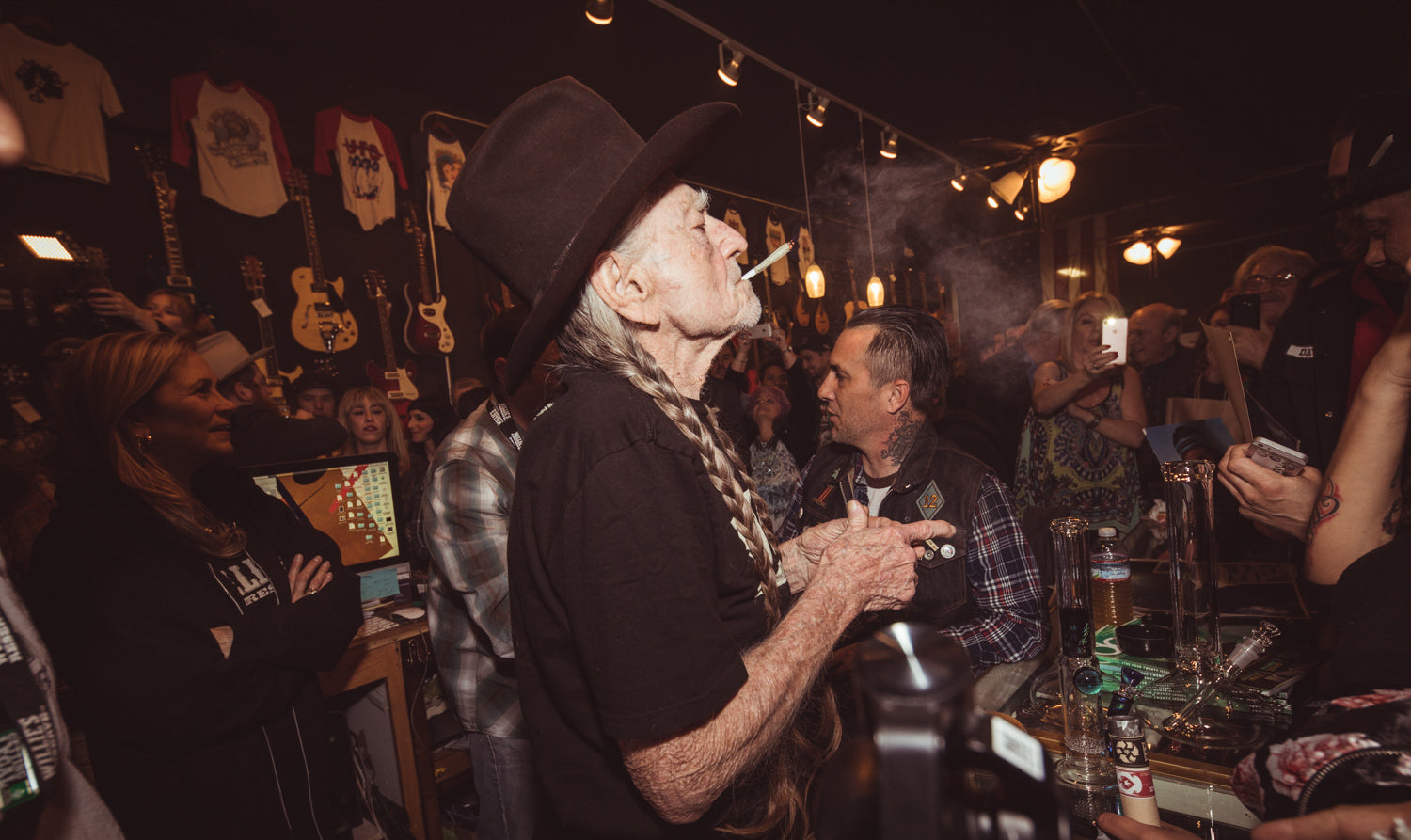 Willie Nelson in a cowboy hat smoking a joint in a lively bar setting with people and guitars in the background.