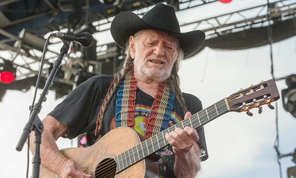 Willie Nelson playing guitar on stage with a microphone and colorful guitar strap.