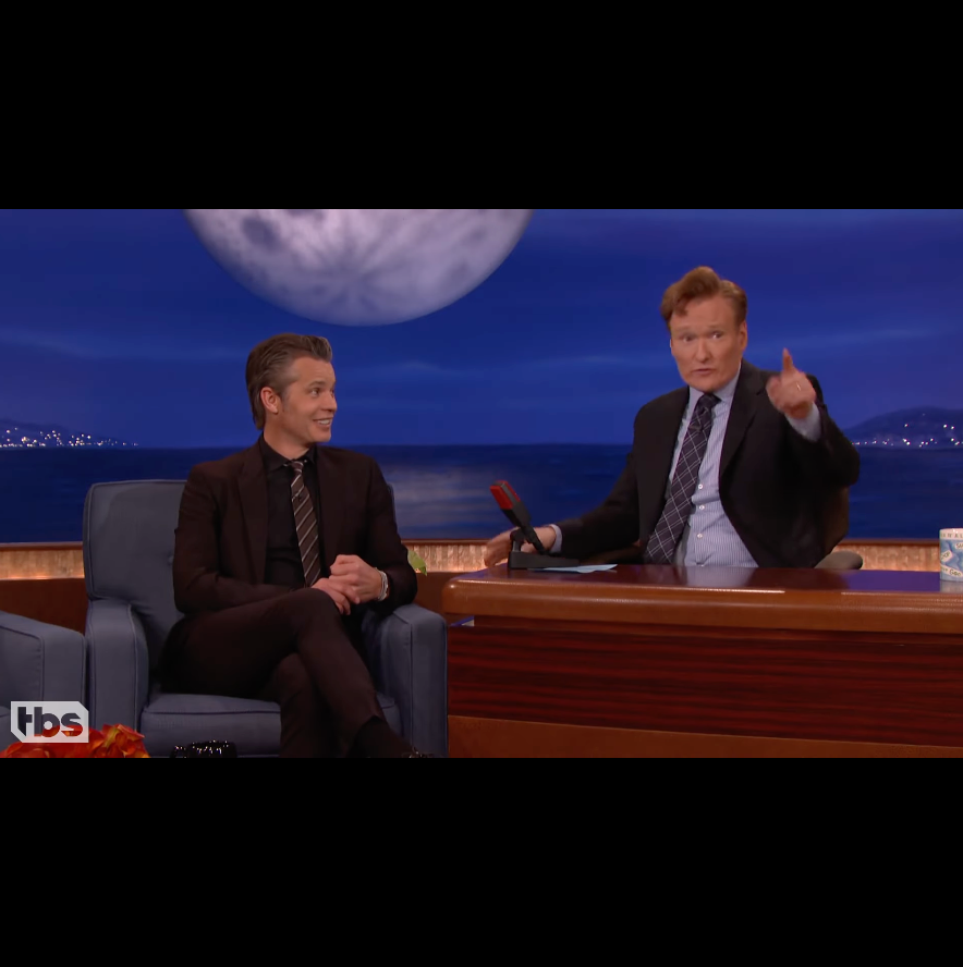 Timothy Olyphant on a talk show set with a moon and night sky background