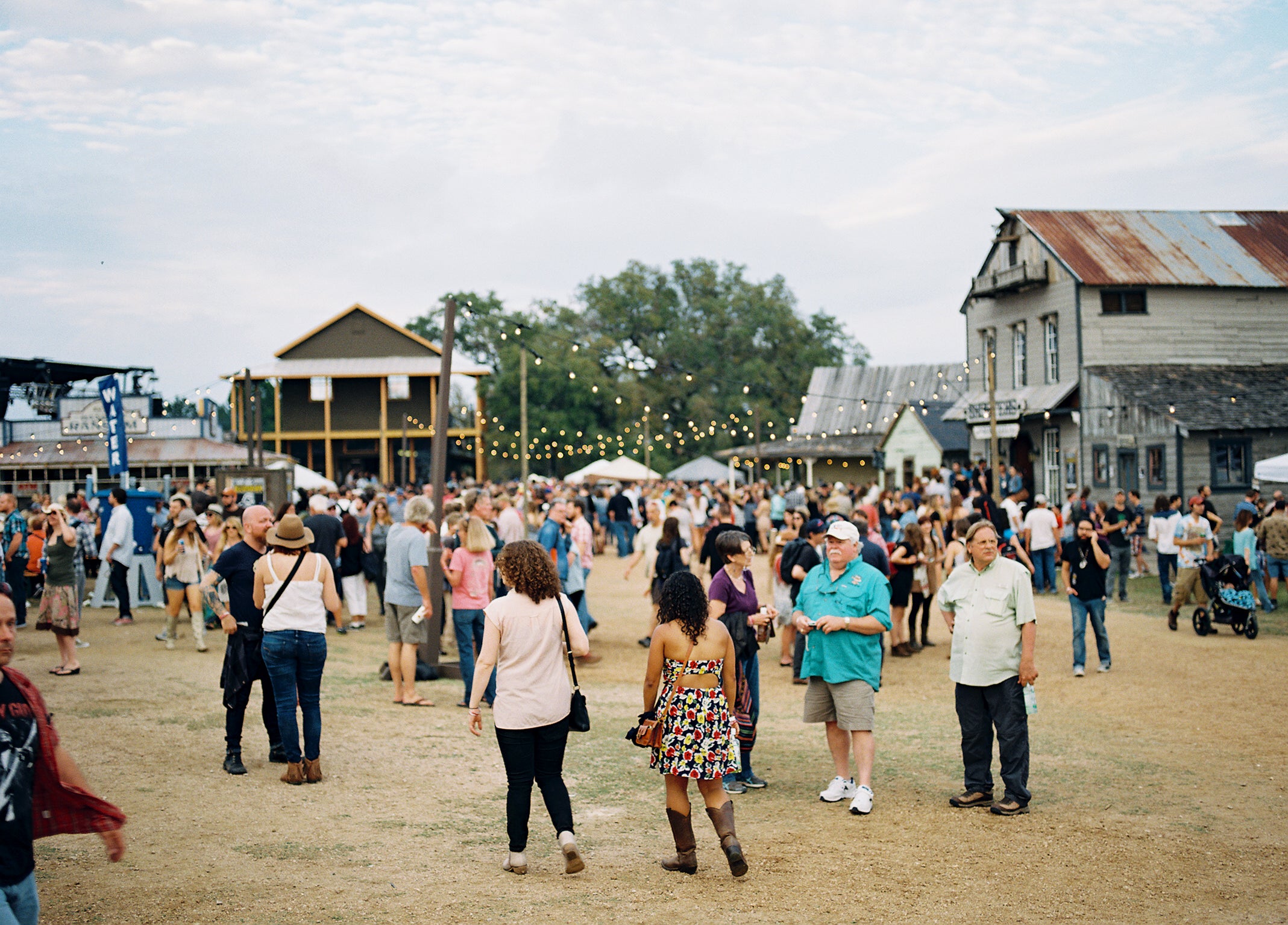 People walking around an outdoor event with wooden buildings and trees in the background.