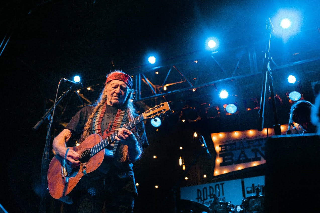 Willie Nelson playing guitar on stage with blue lights and a band name sign in the background