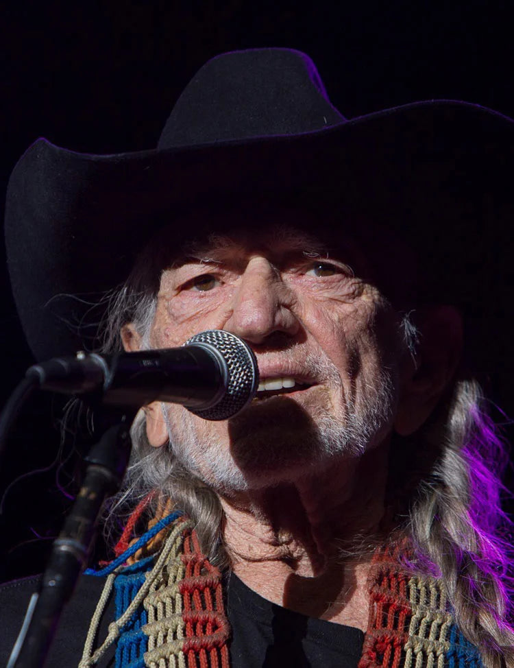 Willie Nelson wearing a cowboy hat and  singing into a microphone with a dark background