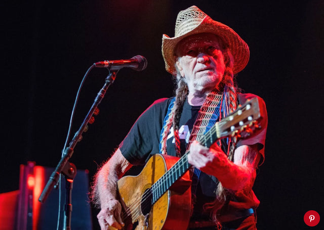Willie Nelson playing guitar on stage with a microphone and dark background