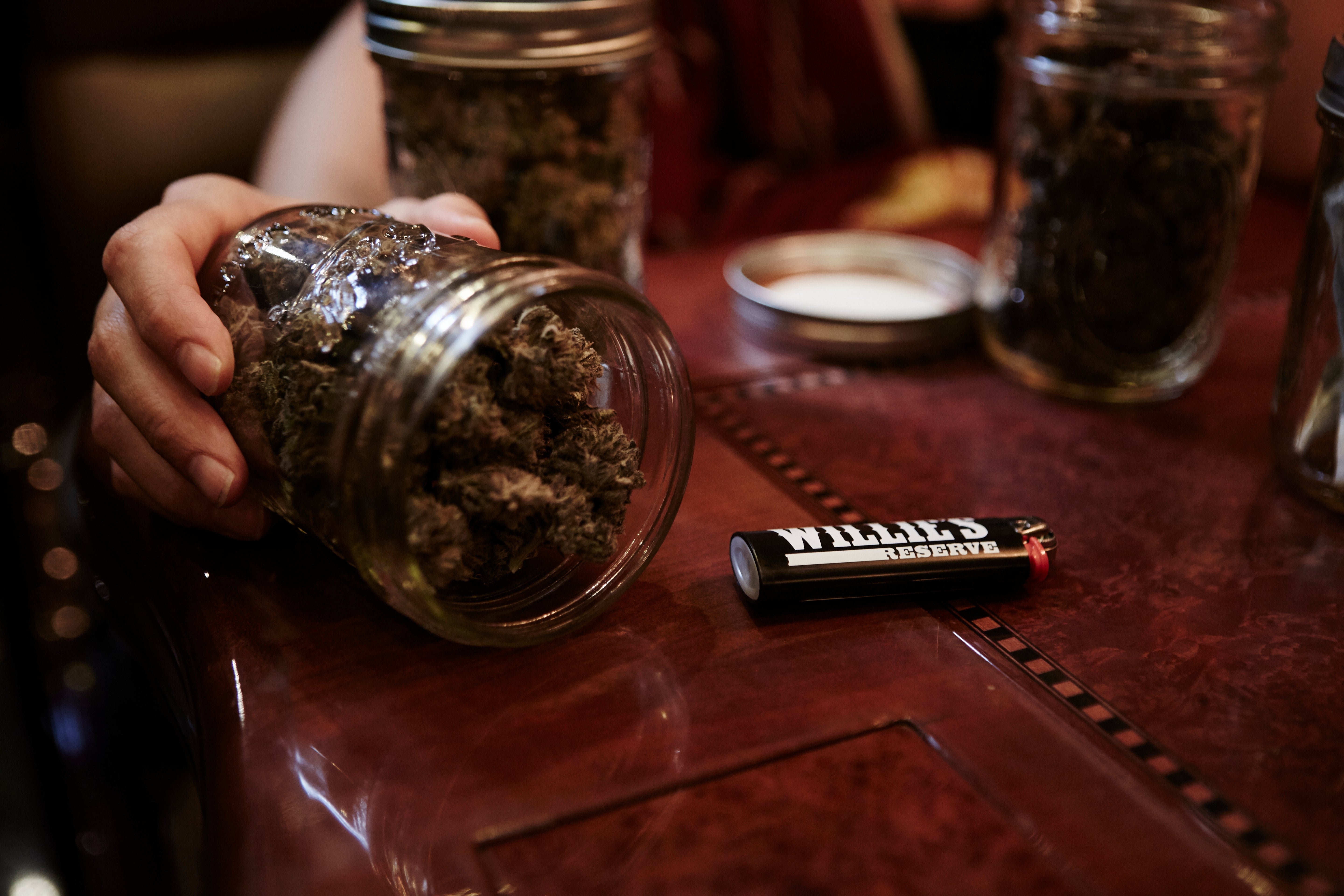 Hand holding a jar of cannabis buds on a table with other jars and a lighter.