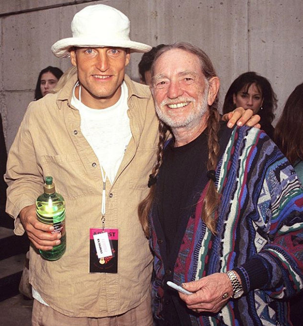 Two men standing together, one holding a bottle, with a concrete wall background