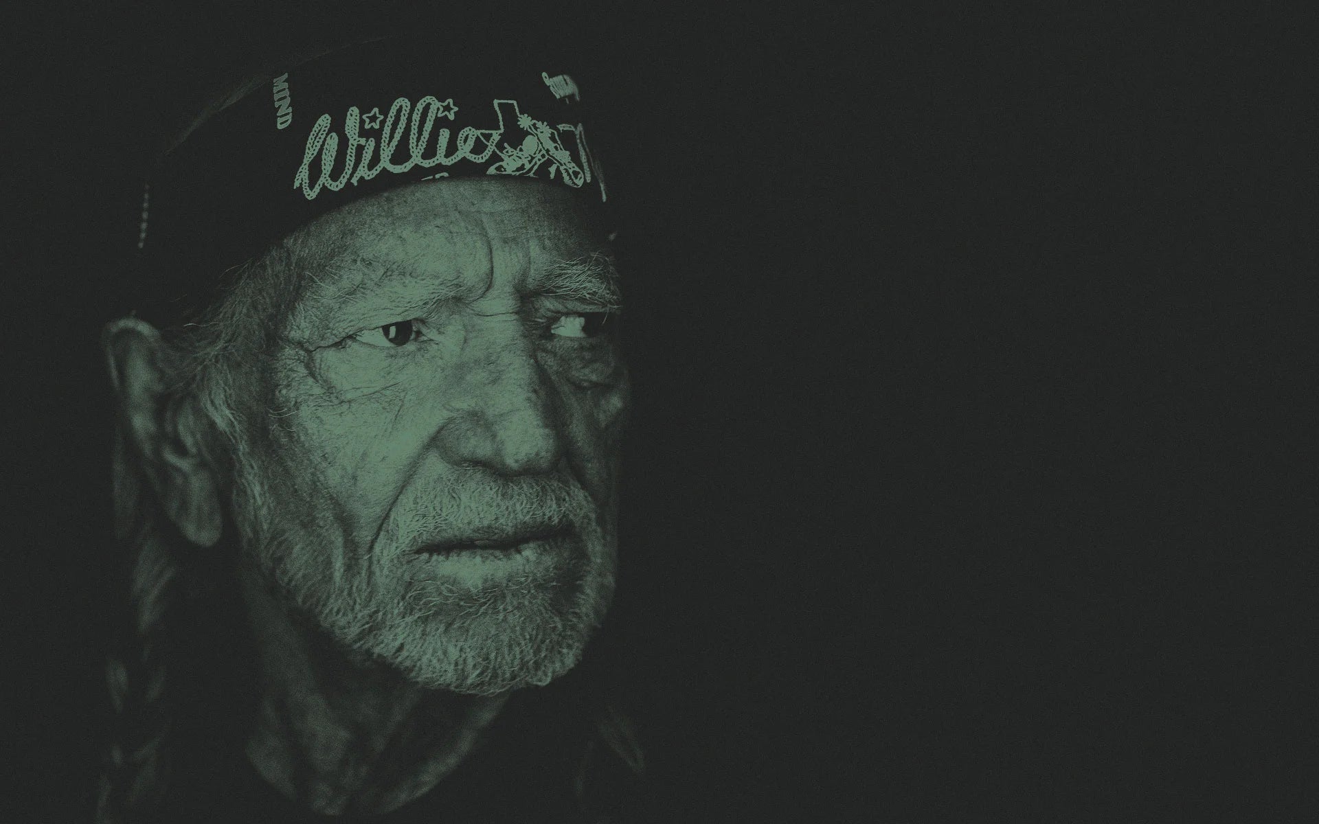 Willie Nelson wearing a cap with 'Willie' on it against a dark background